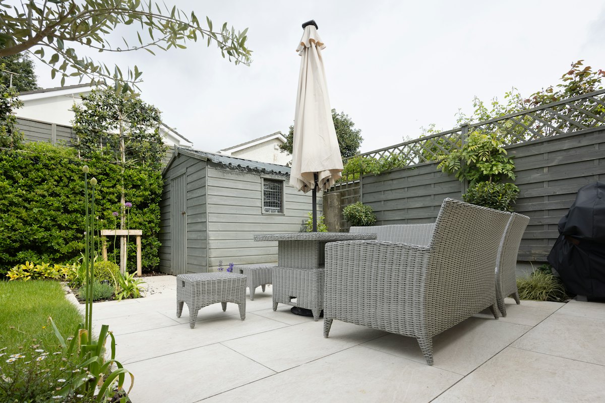 Een terras in grote lichte tegels met tuinmeubels, een parasol en een tuinhuis